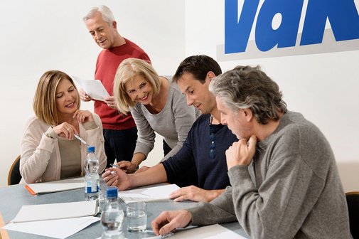 Verschiedene Menschen sitzen an einem Tisch, offenbar in einer Arbeitssitzung, an der Wand hinter ihnen ist ein großes VdK-Logo.