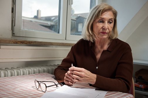 Eine ältere Frau sitzt an einem Küchentisch, sie sieht besorgt und niedergeschlagen aus. Auf dem Tisch vor ihr liegt ein Brief, daneben ihre Lesebrille. 