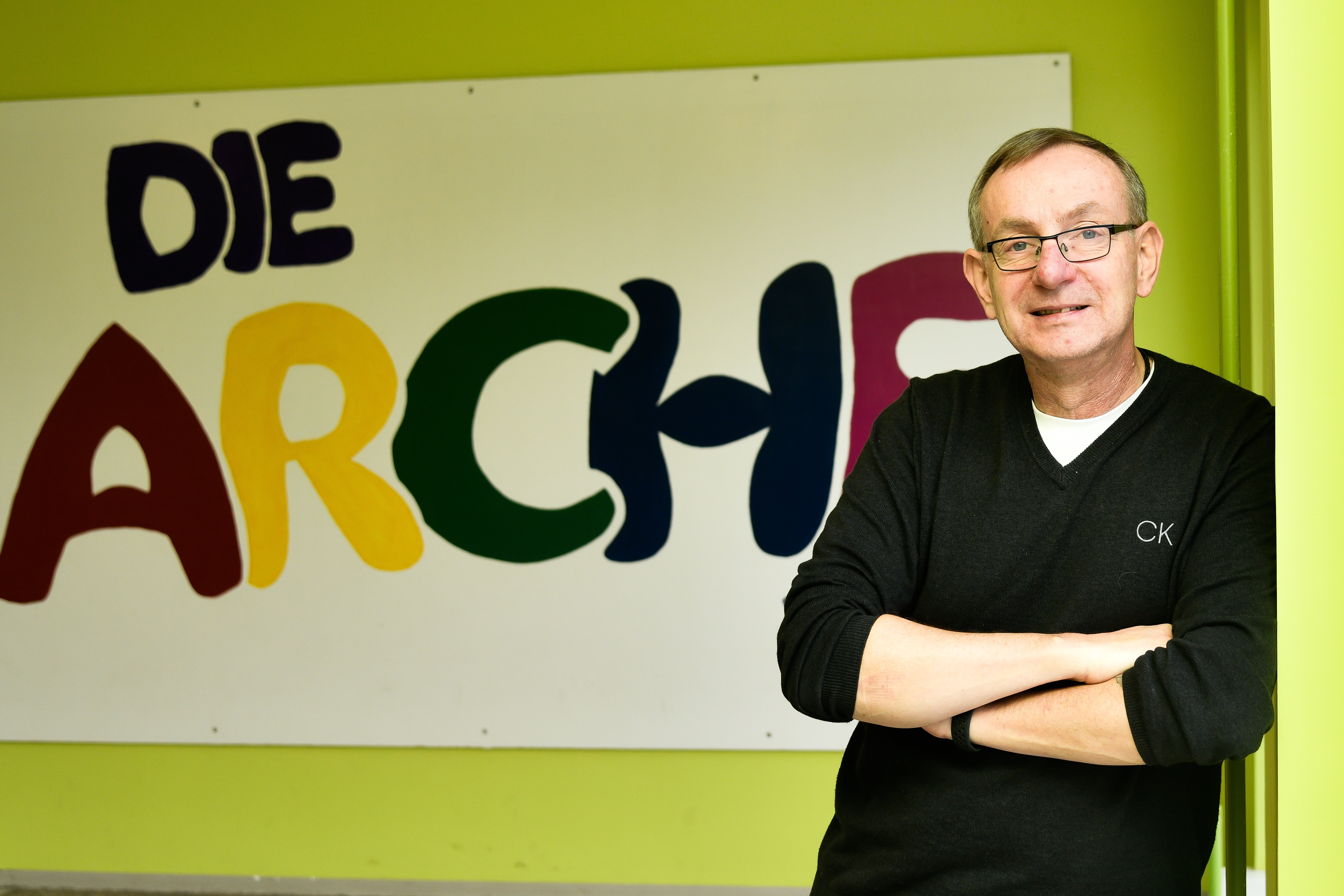 Arche-Gründer Bernd Siggelkow Bernd Siggelkow steht an eine Wand gelehnt, im Hintergrund sieht man ein großes Plakat mit der Aufschrift "Die Arche"