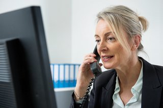 Eine Frau am Computer, sie telefoniert. Im Hintergrund sieht man Aktenordner.