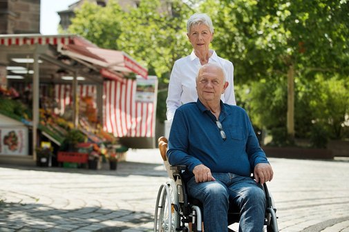 Älteres Paar draußen unterwegs, er sitzt im Rollstuhl, sie schiebt ihn