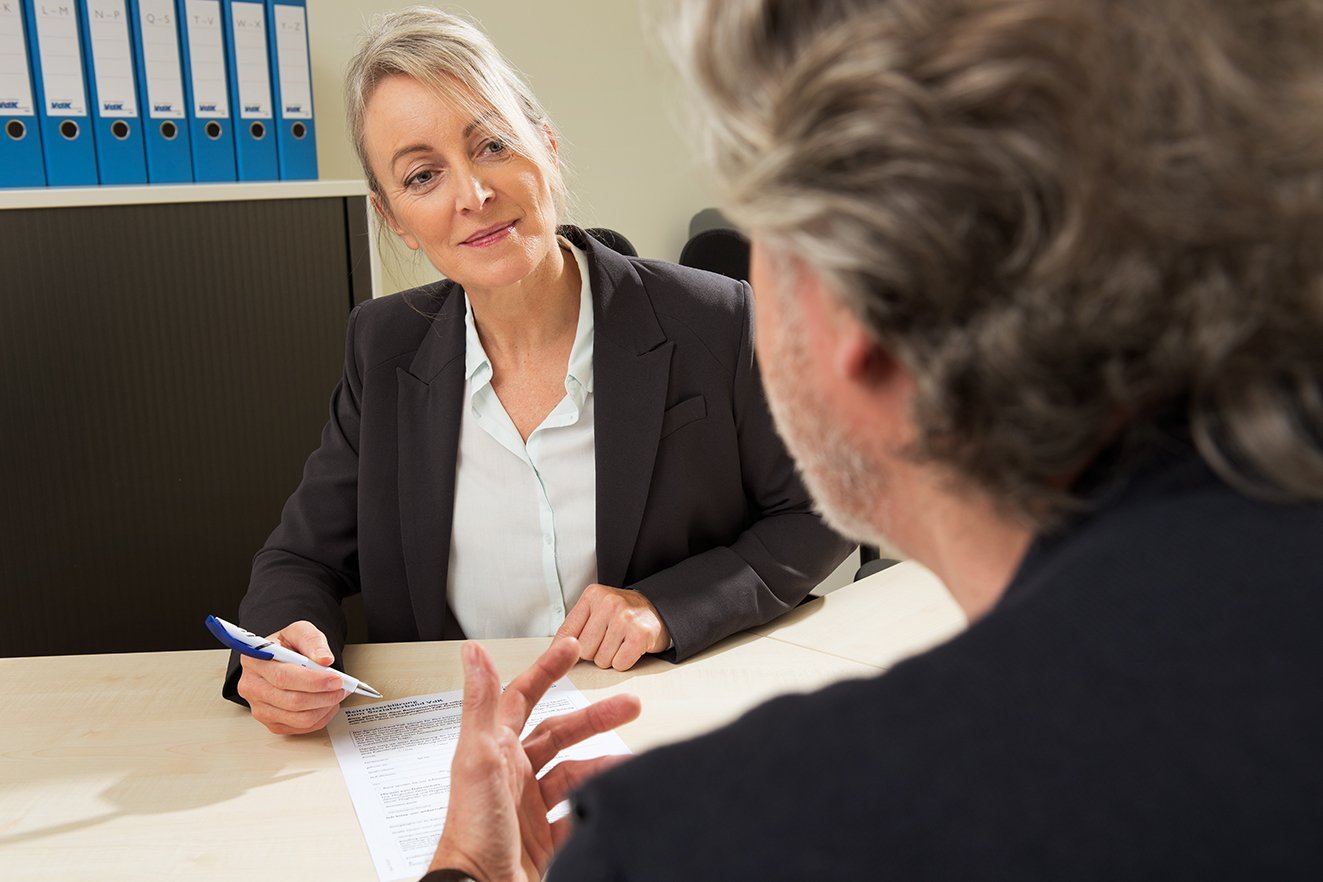 VdK-Beratung Eine Frau reicht einem Mann eine VdK-Beitrittserklärung. Sie sitzen sich in einem Büro gegenüber, im Hintergrund stehen viele Aktenordner im Regal.