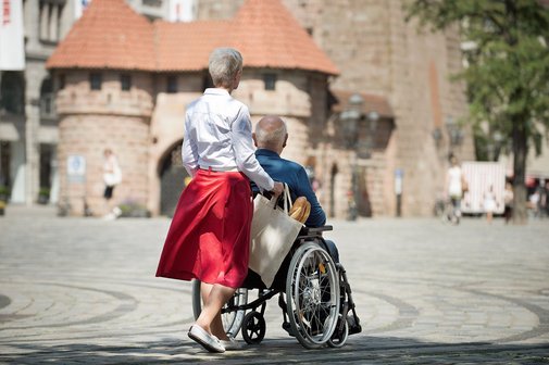 Eine Seniorin schiebt ihren Mann, der im Rollstuhl sitzt, &uuml;ber einen Platz, man sieht im Hintergrund ein altes Stadttor.