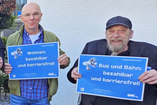 Zwei Ehrenamtliche des VdK halten Plakate mit Forderungen hoch