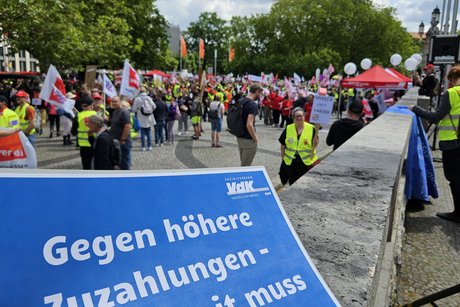 VdK-Plakat mit Demonstranten im Hintergrund