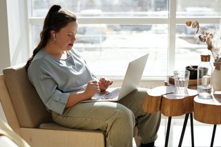 Eine junge Frau mit Down-Syndrom sitzt auf einem Sessel und arbeitet am Laptop