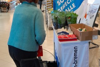 Eine Frau mit Rollator am blau-weißen Infostand. Dahinter sieht man den Verbrauchermarkt
