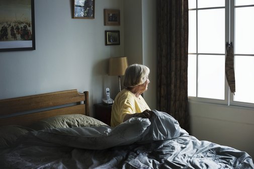Eine &auml;ltere Frau sitzt auf der Kante ihres Bettes und schaut aus dem Fenster