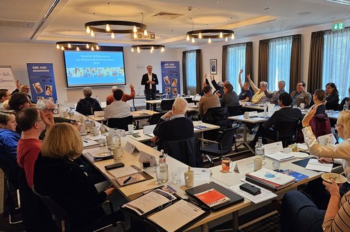Die Teilnehmerinnen und Teilnehmer an Tischen im Saal, ein Teil zeigt auf