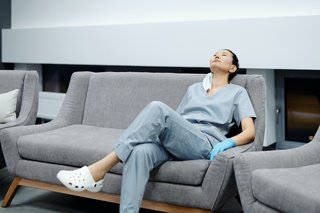 Eine Ärztin in blauer Arbeitskleidung sitzt erschöft auf einem Sofa