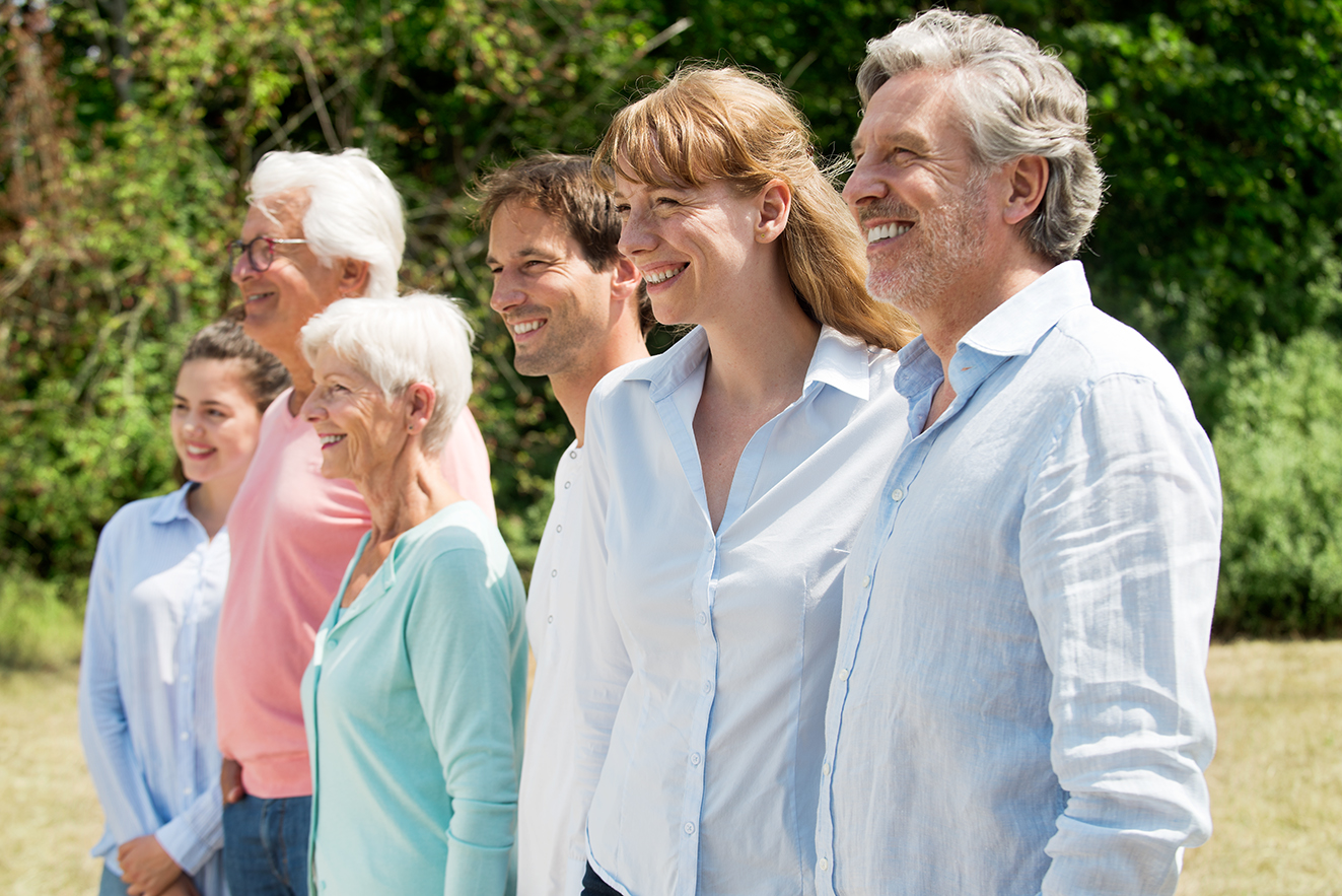 Eine Gruppe aus jungen und alten M&auml;nnern und Frauen stehen nebeneinander, blicken l&auml;chelnd in die Ferne. 