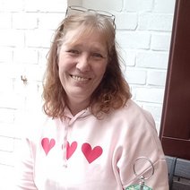 alt="Eine l&auml;chelnde Frau mit r&ouml;tlich-blonden Haaren tr&auml;gt einen hellrosa Kapuzenpullover mit roten Herzen vor einer wei&szlig;en Backsteinwand.