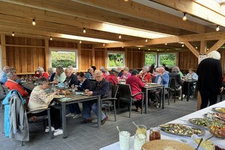 Im Vordergrund des Bildes ist ein reich gedecktes Buffet zu sehen, auf dem verschiedene Salate, Dips und Brotscheiben ansprechend angerichtet sind. Die Tische sind mit Getränken, Tellern und Besteck gedeckt, und es herrscht eine fröhliche, entspannte Stimmung.