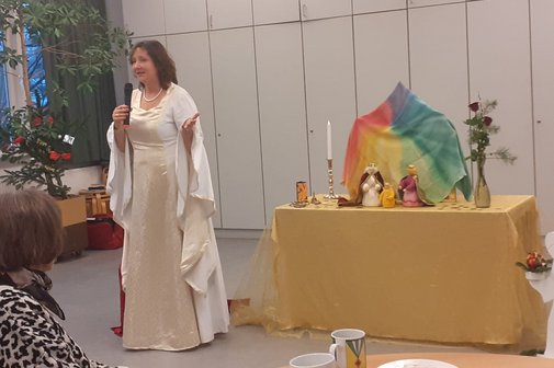 Eine als Christkind verkleidete Frau erz&auml;hlt eine Geschichte. Sie tr&auml;gt ein wei&szlig;es Kleid und steht links neben eine Holzkrippe.