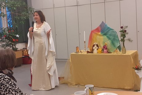 Eine als Christkind verkleidete Frau erz&auml;hlt eine Geschichte. Sie tr&auml;gt ein wei&szlig;es Kleid und steht links neben eine Holzkrippe.