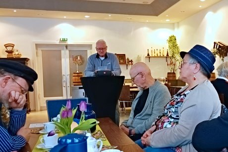 Geschichten auf Plattdeutsch Dieter Gleiß steht hinter einem Stehtisch und liest eine kurze Geschichte auf Plattdeutsch vor. An den Kaffeetischen sitzen die Gäste und hören konzentriert zu.