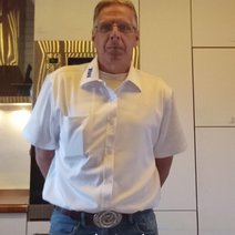 Jan Schweinberger, der zweite Vorsitzende des VdK-Ortsverbands, im offiziellen wei&szlig;en VdK-Kurzarmhemd mit blauem Logo auf dem Kragen. Der Mann mittleren Alters tr&auml;gt eine Brille, blaue Jeans mit einer gro&szlig;en G&uuml;rtelschnalle und blickt direkt in die Kamera. Er steht mit hinter dem R&uuml;cken verschr&auml;nkten Armen vor einer hellen, modernen Wand