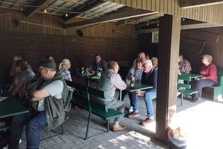 Das Bild zeigt eine Gruppe von Menschen, die an Tischen unter einem hölzernen Unterstand sitzen. Das Treffen befindet sich im Garten vom Alten Fahnster Krug.