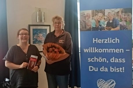 Auf der linken Seite des Bildes steht die Frauenvertreterin des VdK Celle-Altstadt, Bernadette Wolski. Sie hält einen Flyer des Deutschen Roten Kreuzes in der Hand. In der Bildmitte steht Frau Christiane Gentzmann vom DRK. Sie hält den Blumenstrauß in den Händen, der ihr zum Dank für ihren Vortrag überreicht wurde. Auf der rechten Bildseite steht der Rollup des VdK mit der Aufschrift "Herzlich willkommen - schön, dass Du da bist!"