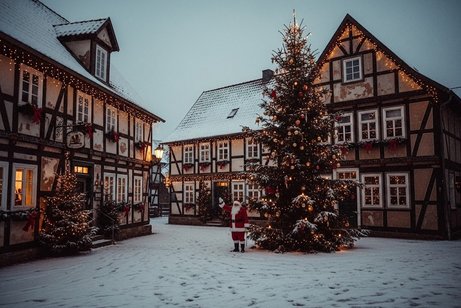 Niedersächsische Fachwerkhäuser, vor denen ein großer Weihnachtsbaum steht. Neben dem Baum steht der Weihnachtsmann und winkt. Es liegt Schnee.