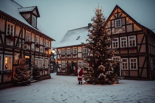 Niedersächsische Fachwerkhäuser, vor denen ein großer Weihnachtsbaum steht. Neben dem Baum steht der Weihnachtsmann und winkt. Es liegt Schnee.