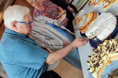 Auf einem Tisch stehen Torten und Kuchen. Einige Teilnehmer bedienen sich.