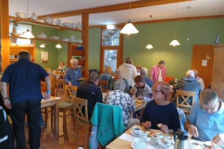 Eine Innenaufnahme des Heide-Hauses, gef&uuml;llt mit G&auml;sten des VdK-Sommerfests, die an hellen Holztischen sitzen, Kaffee trinken und essen. Die gr&uuml;nen W&auml;nde sind mit Holzbalken und Regalen verziert, auf denen wei&szlig;e Keramik steht, was eine gem&uuml;tliche Atmosph&auml;re schafft. Menschen unterhalten sich, darunter &auml;ltere Herrschaften und ein Mann in einem blauen Hemd, der im Vordergrund sitzt. Das Personal des Hauses scheint den Service zu organisieren. Der Raum ist hell und durch die gro&szlig;en Fenster im Hintergrund sichtbar. Eine T&uuml;r f&uuml;hrt auf eine Terrasse. Die Szene vermittelt ein Gef&uuml;hl von Gemeinschaft und Freude.