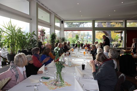 Das Foto zeigt mehrere Personen, die an einer langen Tischreihe sitzen. Auf den Tischen liegen weiße Tischdecken und herbstliche Dekoration.