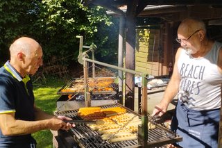 Das Bild zeigt zwei Personen, die im Freien an einem Grill stehen. Auf dem Grill befinden sich Würstchen und Grillfleisch. Die Umgebung ist der Garten vom Alten Fahnster Krug.