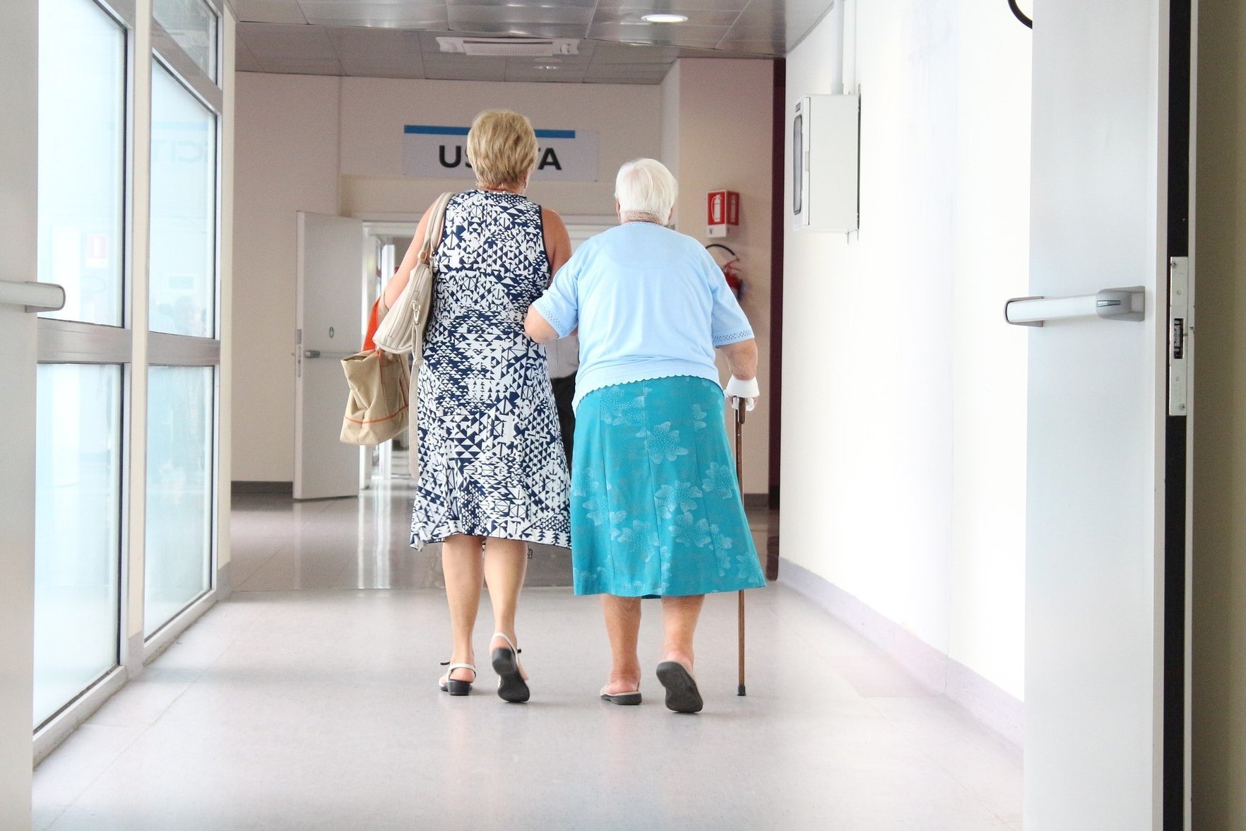 Eine Frau geht mit einer &auml;lteren Frau mit Gehstock einen Krankenhausflur entlang