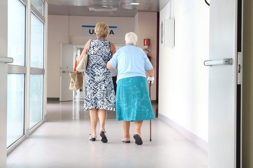 Eine Frau geht mit einer &auml;lteren Frau mit Gehstock einen Krankenhausflur entlang