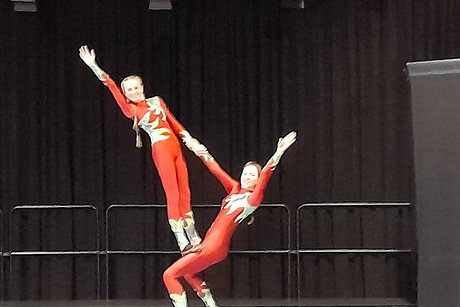 Zwei junge Artistinnen präsentieren eine Hebeübung, bei der ein Mädchen auf dem Oberschenkel des zweiten Mädchens steht und sich gegenseitig mit einer Hand halten. Der freie Arm wird dabei nach außen geschwenkt