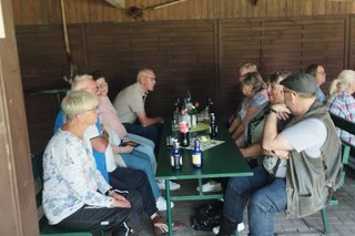 Das Bild zeigt eine Gruppe von Menschen, die an Tischen unter einem hölzernen Unterstand sitzen. Das Treffen befindet sich im Garten vom Alten Fahnster Krug.