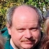 Ein Mann mit Geheimratsecken und sch&uuml;tterem, grauem Haar, der einen Schnurrbart tr&auml;gt und vor einer gr&uuml;nen Hecke steht.