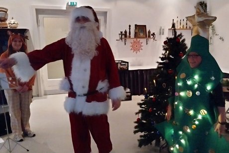 Der Weihnachtsmann tr&auml;gt ein Gedicht vor. Neben ihm stehen ein singendes Rentier und ein klavierspielender Tannenbaum.