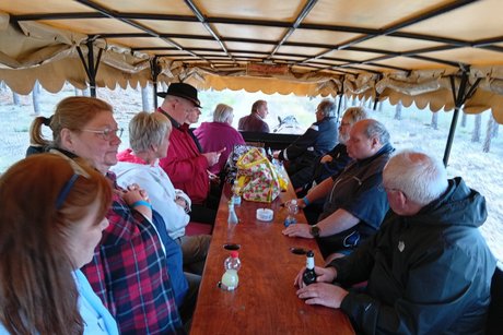 Eine Gruppe von etwa zehn Personen sitzt an einem langen Holztisch im Innenraum eines Planwagens. Die Sicht geht in Fahrtrichtung, w&auml;hrend die Gruppe durch eine Heide- und Waldlandschaft f&auml;hrt. Die Fahrg&auml;ste, darunter &auml;ltere Menschen, sind in Freizeitkleidung und unterhalten sich angeregt. Ein Kutscher lenkt das Gespann. Die Szene vermittelt ein Gef&uuml;hl der Geselligkeit und Ausflugsstimmung