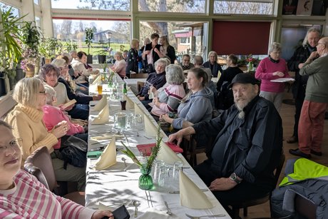 Das Foto zeigt mehrere Personen, die an einer langen Tischreihe sitzen. Auf den Tischen liegen wei&szlig;e Tischdecken und fr&uuml;hlingshafte Dekoration.