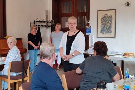 Frau Müller steht vor den Tisvhen mit den Anwesenden.