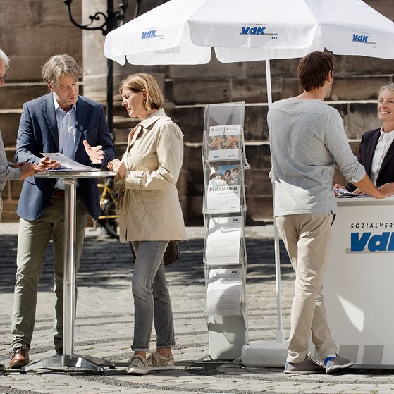 Ein VdK-Infostand in einer Fu&szlig;g&auml;ngerzone, eine Frau spricht mit einem Passanten, am Stehtisch daneben unterhalten sich Menschen. 
