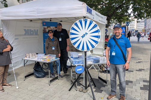 Der VdK-Stand am Platz der Weltausstellung Auf dem Bild ist ein VdK-Zeltstand zu sehen. Darin stehen zwei Personen. Links davor steht eine weibliche Person mit Blindenstock. Rechts daneben steht ein Glücksrad und eine männliche Person mit blauer Sonnenkappe und blauem Polohemd.