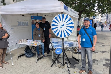 Auf dem Bild ist ein VdK-Zeltstand zu sehen. Darin stehen zwei Personen. Links davor steht eine weibliche Person mit Blindenstock. Rechts daneben steht ein Gl&uuml;cksrad und eine m&auml;nnliche Person mit blauer Sonnenkappe und blauem Polohemd.