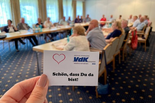 Eine Karte mit "Sch&ouml;n, dass du da bist". Im Hintergrund VdKler bei einem Treffen an Tischen
