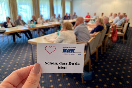 Eine Karte mit "Sch&ouml;n, dass du da bist". Im Hintergrund VdKler bei einem Treffen an Tischen