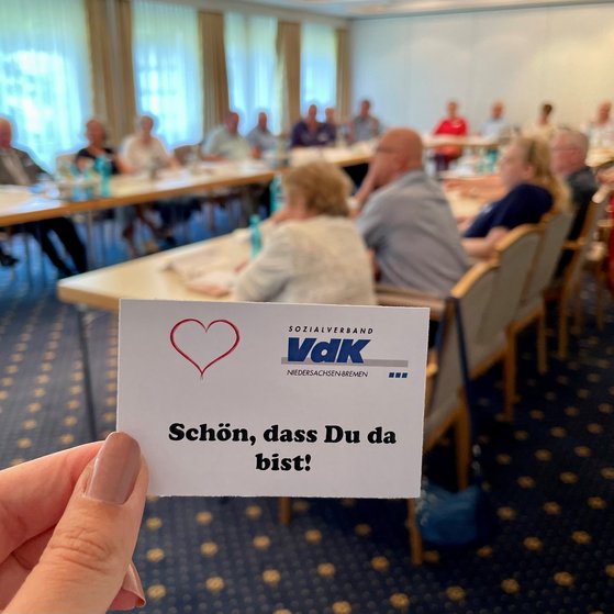 Eine Karte mit "Sch&ouml;n, dass du da bist". Im Hintergrund VdKler bei einem Treffen an Tischen