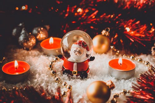 Eine Schneekugel mit Weihnachtsmann-Figur steht inmitten von Kunstschnee, brennenden Teelichtern und goldenen Christbaumkugeln