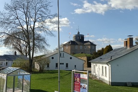 Bei sonnigem Wetter sind die Bauten (Unterkünfte, Küche etc.) in der Grünanlage sehr gut im Vordergrund zu erkennen. Im Hintergrund hebt sich auf einem grünen Hügel die Festung Wilhelmstein ab.