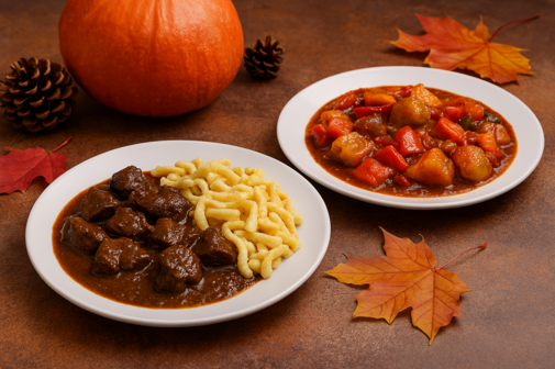 Ein Bild mit zwei Tellern: Einer enthält Hirschgulasch und Spätzle, der andere vetarisches Gulasch. Beide stehen vor einem herbstlichen Hintergrund.