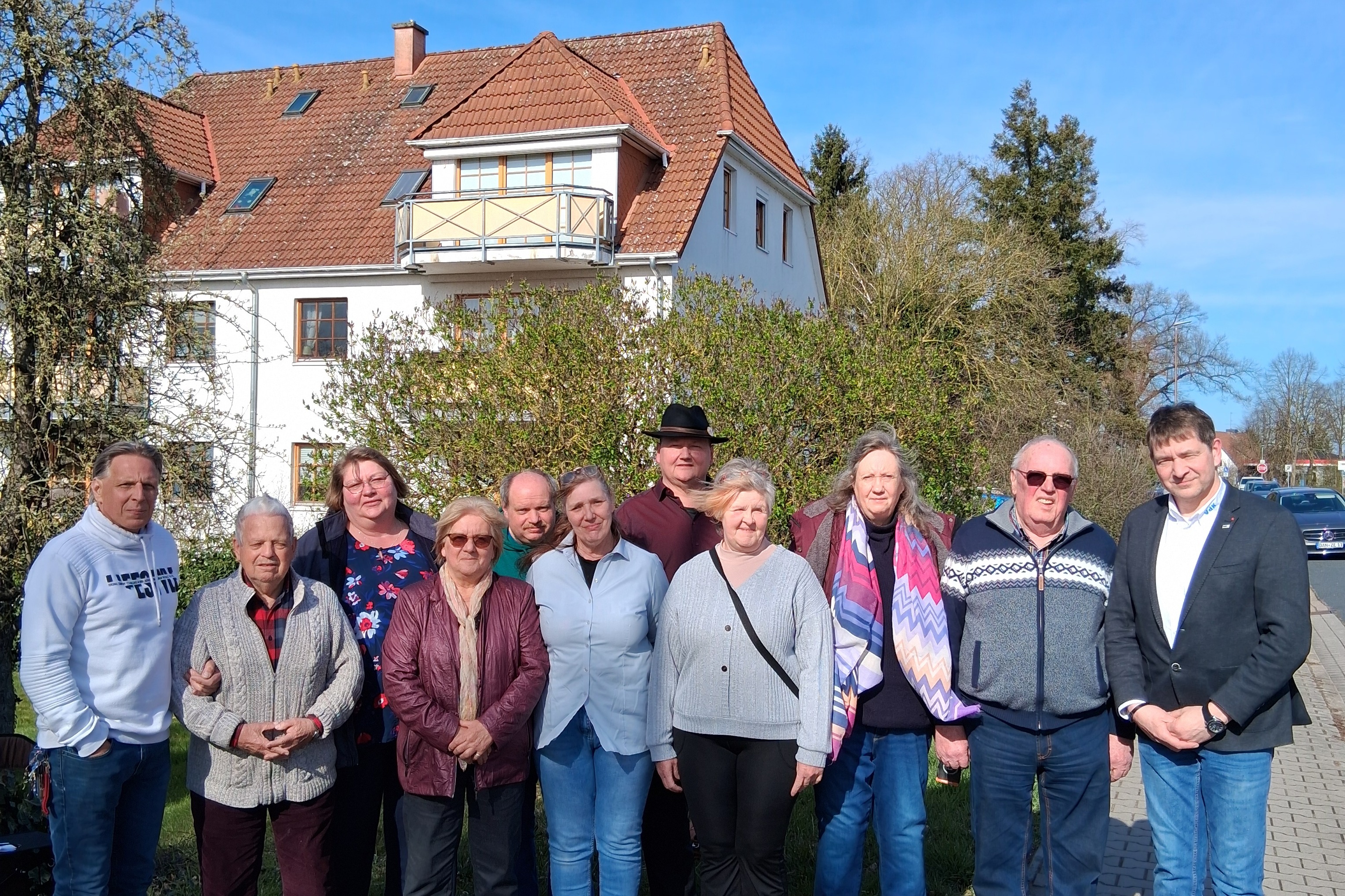 Eine Gruppe von elf Personen steht im Freien f&uuml;r ein Gruppenfoto nebeneinander auf einem Gehweg an einer Stra&szlig;e. Im Hintergrund ist ein zweist&ouml;ckiges Wohnhaus mit hellem Putz, rot gedecktem Dach und Balkon zu sehen, umgeben von B&auml;umen und Str&auml;uchern bei sonnigem Wetter. Links im Vordergrund steht ein Rollator auf der Rasenfl&auml;che