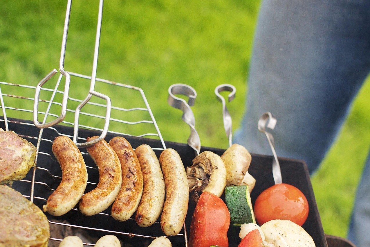 Grill mit Grillgut Eine Person steht am Grill mit einer Grillgabel und bereitet Spieße, Bratwurst und Steaks