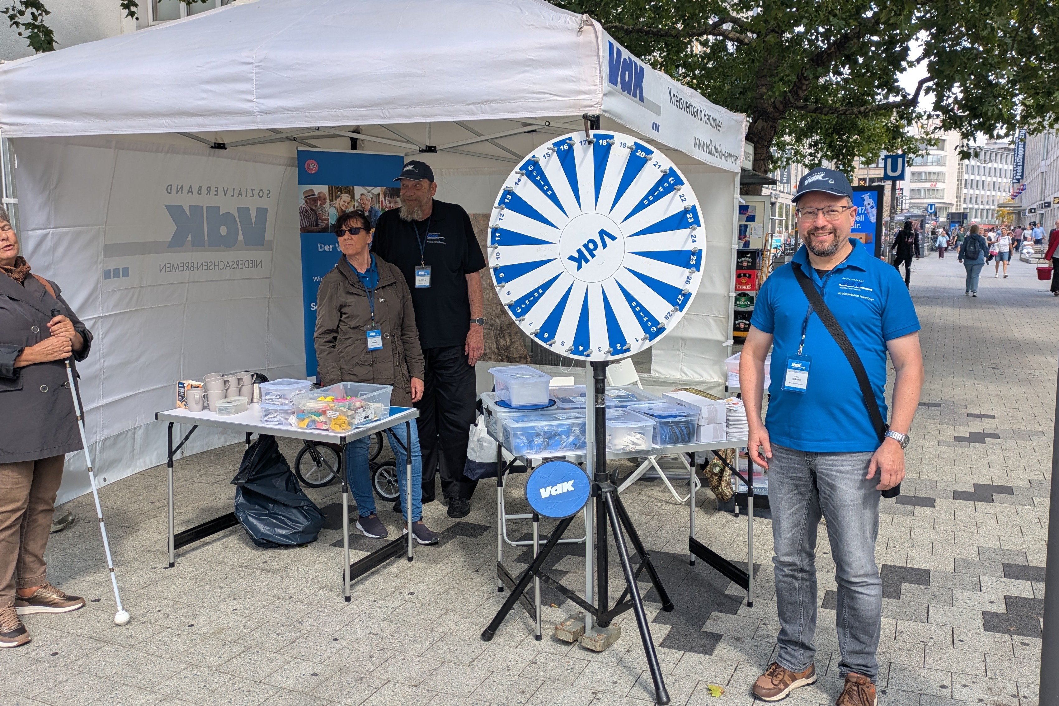 Der VdK-Stand am Platz der Weltausstellung Auf dem Bild ist ein VdK-Zeltstand zu sehen. Darin stehen zwei Personen. Links davor steht eine weibliche Person mit Blindenstock. Rechts daneben steht ein Glücksrad und eine männliche Person mit blauer Sonnenkappe und blauem Polohemd.